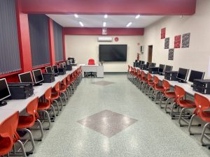 Pracownia komputerowa z czerownymi akcenami na ścianach i suficie. Po obu stronach sali znajdują się 2 rzędy stolików z czerwonymi krzesłami. Na każdym stoliku znajdują się 2 komputery i monitor. Na pczątku sali znajduje się biurko nauczyciela, czerwony fotel i monitor interaktywny.