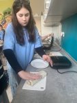Uczennica podczas robienia tortilli na lekcji techniki. W tle opiekacz, sprzęt kuchenny i plakat z warzywami