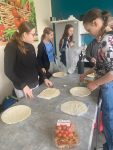 Uczennice podczas robienia tortilli na lekcji techniki. W tle plakat z koszem warzyw i białe meble.