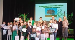 Grupa kolorowo ubranych dzieci z dyplomami w ręce stojąca na scenie na tle wyświetlanego na ekranie zielonego napisu Wojewódzki Konkurs Piosenki Ekologicznej Jak skowronek pod spodem obrazek drzewa