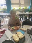 Uczeń w jasnej bluzie siedzi przy szarym stoliku i trzyma w ręce pudełko z warzywami-pomidor, ogórek ,sałata , przed nim na talerzu położone są dwa kawałki bagietki , z tyłu siedzą przy stolikach inni uczniowie w kolorowych ubraniach na tle okien sali lekcyjnej