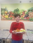 Uczeń w czerwonej bluzie stoi na tle jasnej ściany z fototapetą z kolorowymi owocami i warzywami trzymając w ręce talerz z kolorowymi kanapkami z szynką, ogórkami, pomidorami i rzodkiewką .
