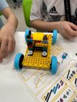Na białej ławce szkolnej stoi pojazd zbudowany z klocków Lego z żółtym podwoziem niebieskimi kółkami w środku którego siedzą dwa ludziki zbudowane z klocków Lego, w tle widnieją ręce dwóch uczniów sterujących pojazdem.