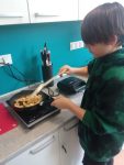 Chłopiec w czarnej bluzie w czarne wzory stoi przy szarej szafce kuchennej smażąc na kuchence indukcyjnej w czarnej patelni pokrojone mięso z kurczaka, na tle niebieskiej ściany stoją na szafce czarne tostery i pojemnik z nożami