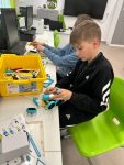 Uczeń w czarnej bluzie siedzi i konstruuje z kolorowych klocków Lego pojazd , montując niebieskie elementy podwozia, przed nim po lewej stronie żółte pudło z elementami klocków, w tle sala lekcyjna z biurkiem i tablicą interaktywną na białej ścianie. Robotyki SkriBot